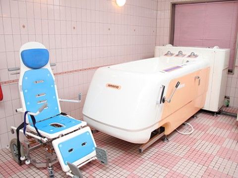 ピンクと白のタイル張りの浴室で、ハンドルの付いた大型の浴槽の手前に、青色の座面の車椅子が置かれている。