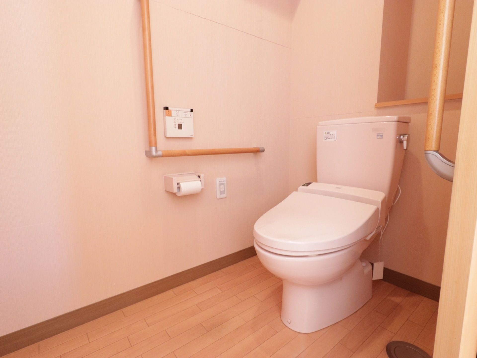 トイレ個室内の様子。洋式便器があり、壁に操作ボタンがある。壁にはＬ字型の手すりとナースコールも設置されている。
