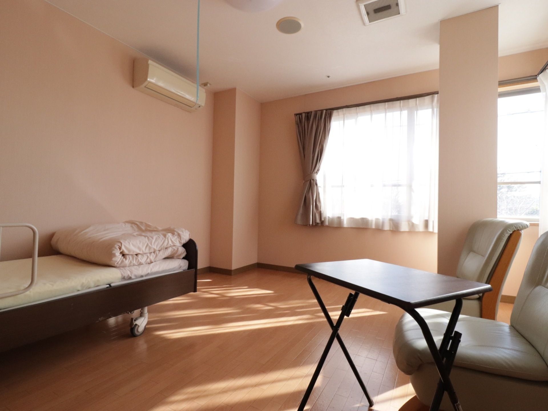 部屋の奥に窓があり、壁にエアコンが設置されている。床は茶色いフローリングである。部屋の一角に横広の柱がある。