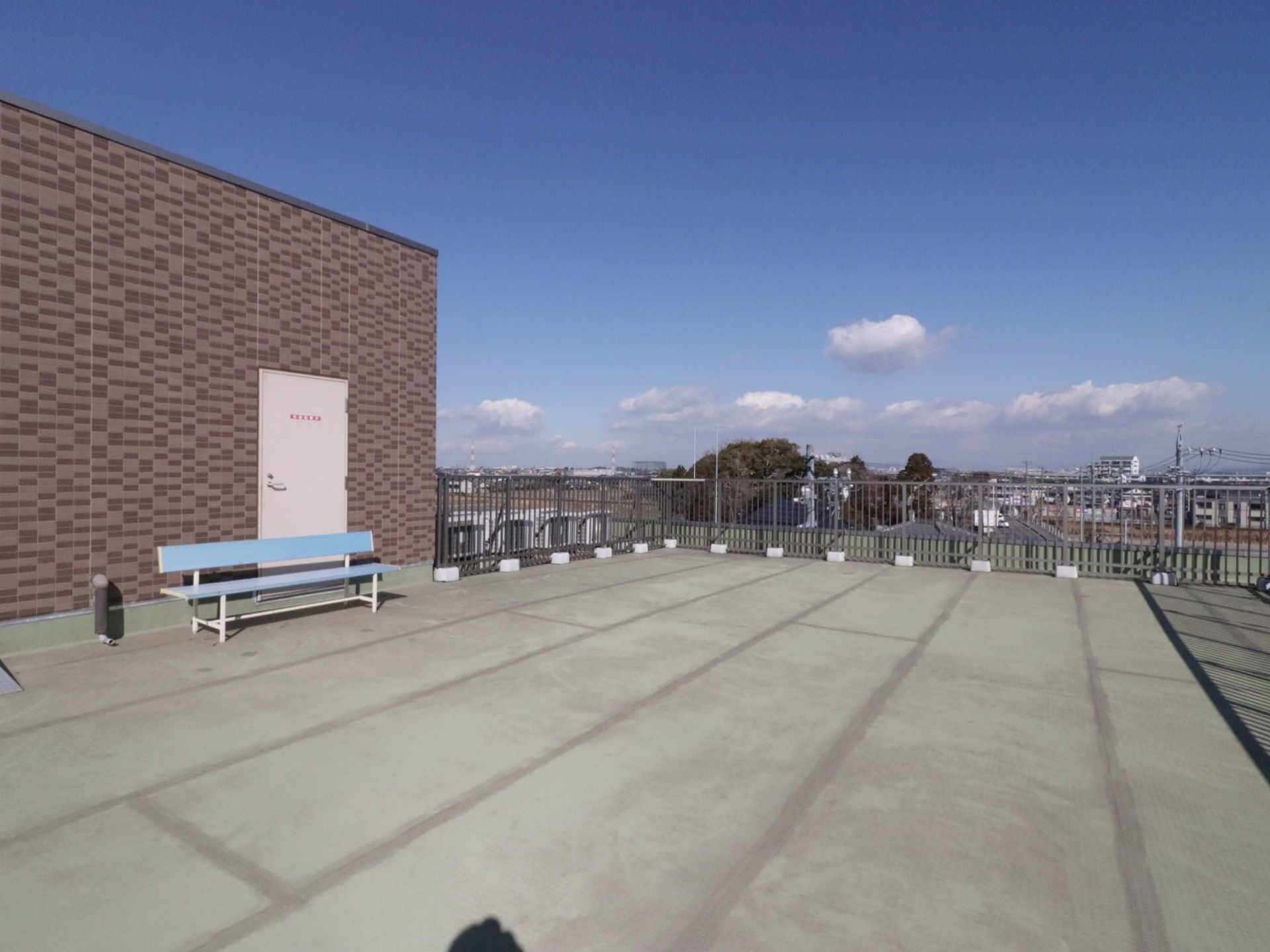 屋上の床は、コンクリート造りである。左手にレンガ風の四角い建物があり、水色のベンチが１台置かれている。