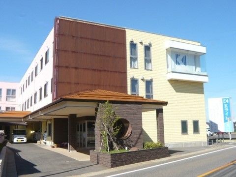 白い外壁の建物に出窓が付いており、一部の壁は茶色になっている。道路の側には、施設名の書かれた水色の看板が立っている。