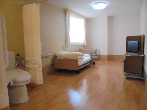電動ベッドの上に布団が畳んでおいてあり、茶色の台にはテレビが設置されている。部屋には仕切りがなく、車椅子をご利用の方でも快適に過ごせる。