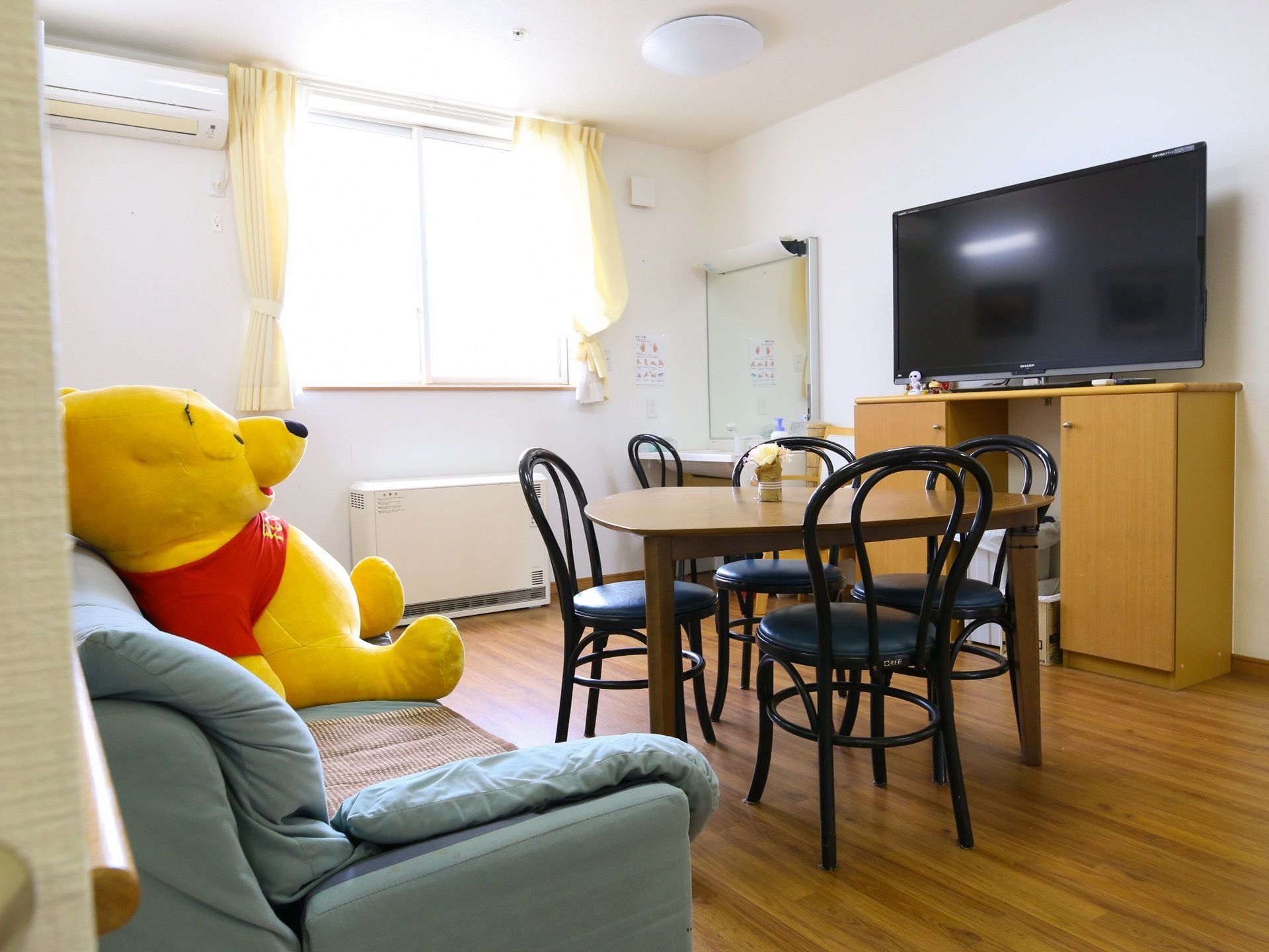 大型のテレビがある部屋には、４脚の椅子を置いたテーブルと、グリーンのソファーが設置されている。角にある洗面台にも椅子がセットしてある。
