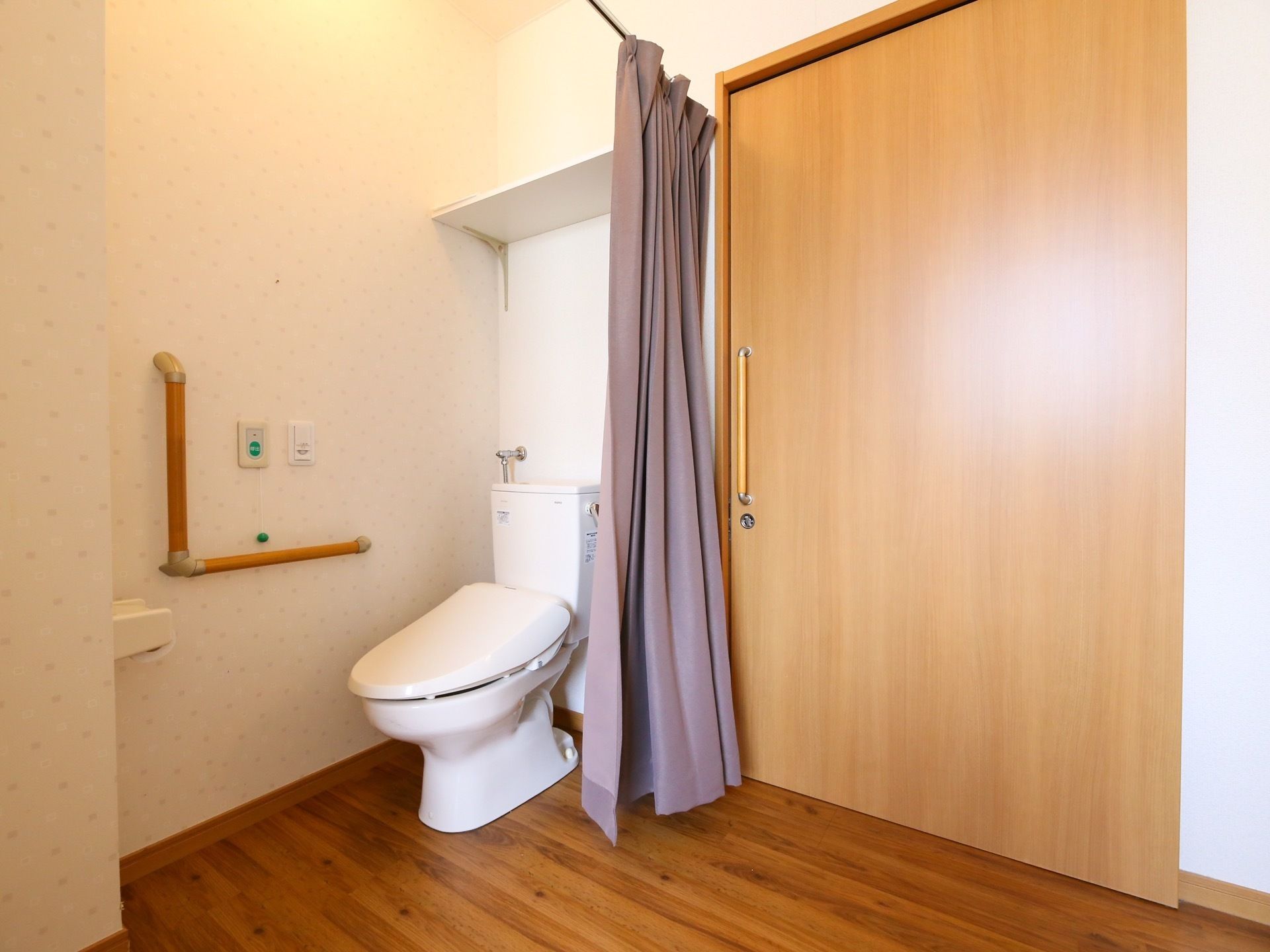 居室のトイレは安全面に配慮したつくりで、ドアがなく、紫色のカーテンがかかっている。壁にはナースコールと手すりがある。
