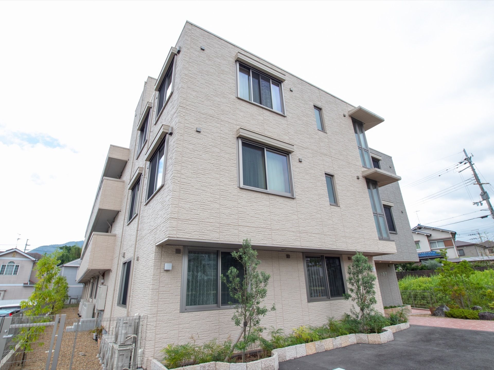 3階建てのベージュ色の建物があり、敷地はアスファルト舗装されている。植栽スペースに樹木が植えられ、周辺には民家が建っている。