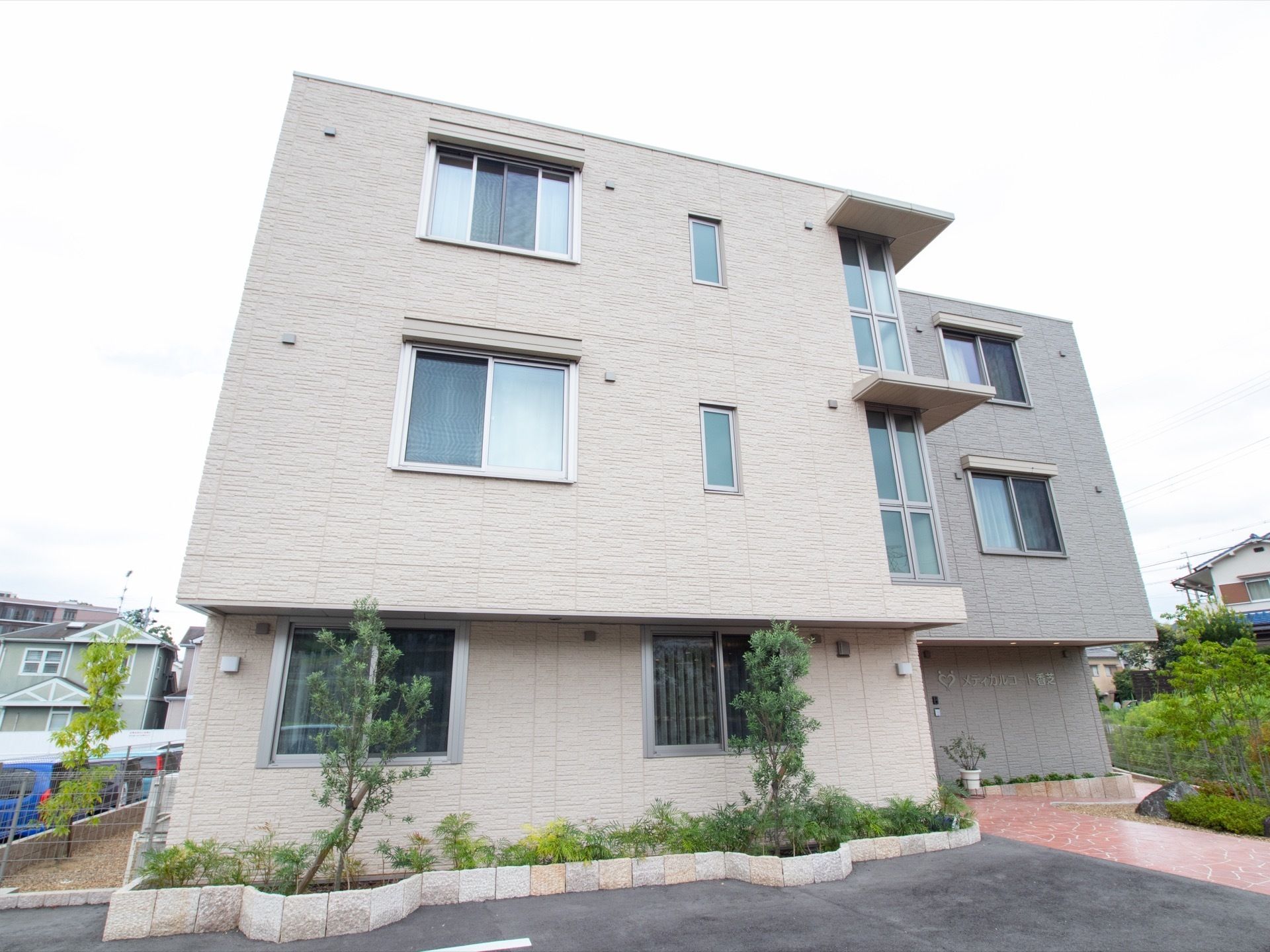 落ち着いたベージュ系の色合いの3階建ての建物である。外壁に施設名の看板が掲げられている。きれいに手入れされた植木がある。