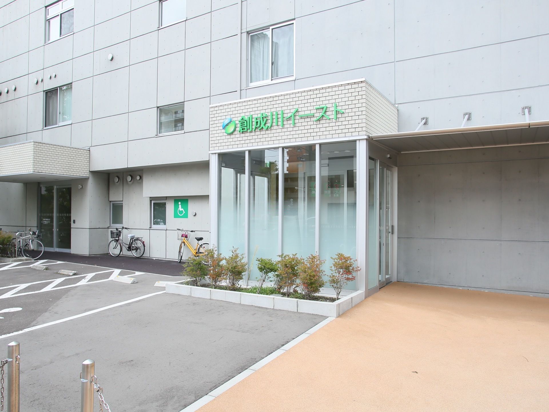 コンクリート壁の建物で、玄関はガラスで囲まれている。上部に明るい緑色でロゴマークと施設名が書かれている。