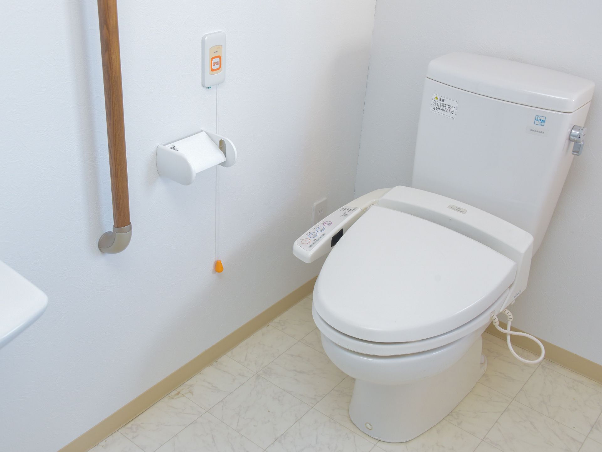 壁に呼び出しブザーが取り付けてある洋式のトイレである。温水洗浄機がついていて、シートの横に操作パネルが取り付けてある。