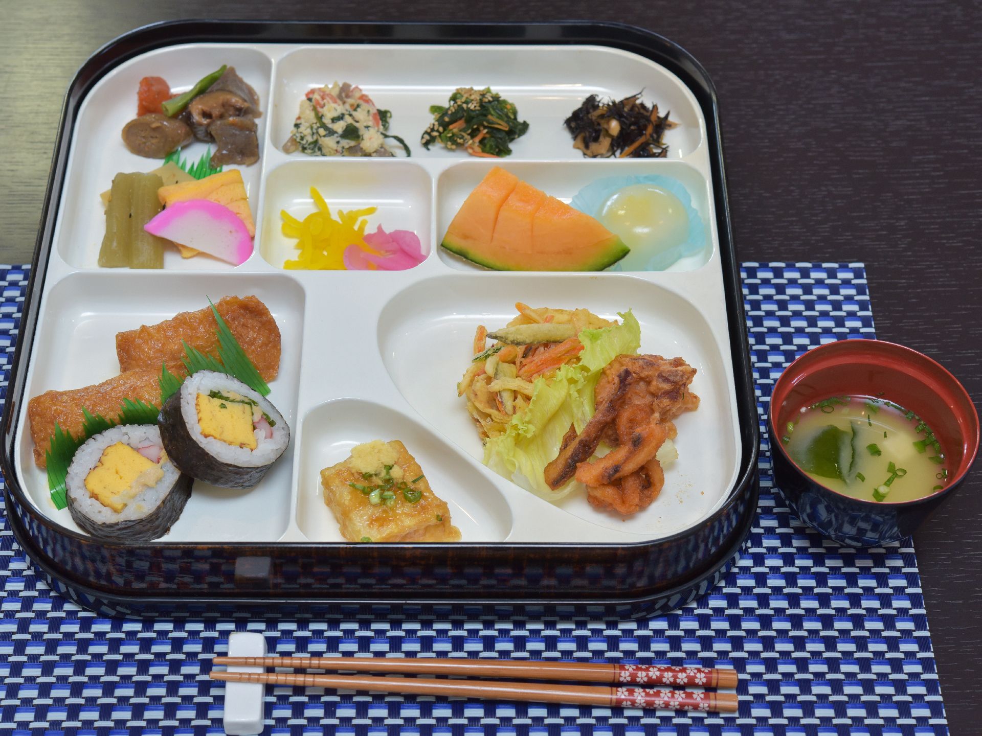 仕切りがついたお皿に、おかすがいろいろと盛ってある。味噌汁がついている。メニューの内容は稲荷、海苔巻き、香の物などである。