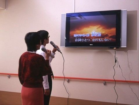 壁に設置されているテレビに流れる歌詞を見て歌う二名の入居者