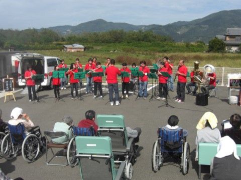 屋外にて、赤いユニフォームを着た楽団が演奏している様子を眺めている、入居者たち