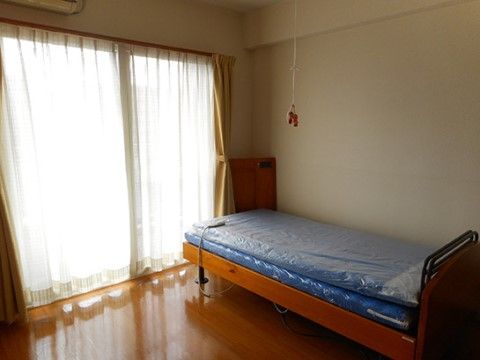 エアコンが完備された個室タイプの居室。壁沿いに青いマットが敷かれたベッドが置かれており、窓にはレースと厚手のカーテンが取り付けられている。