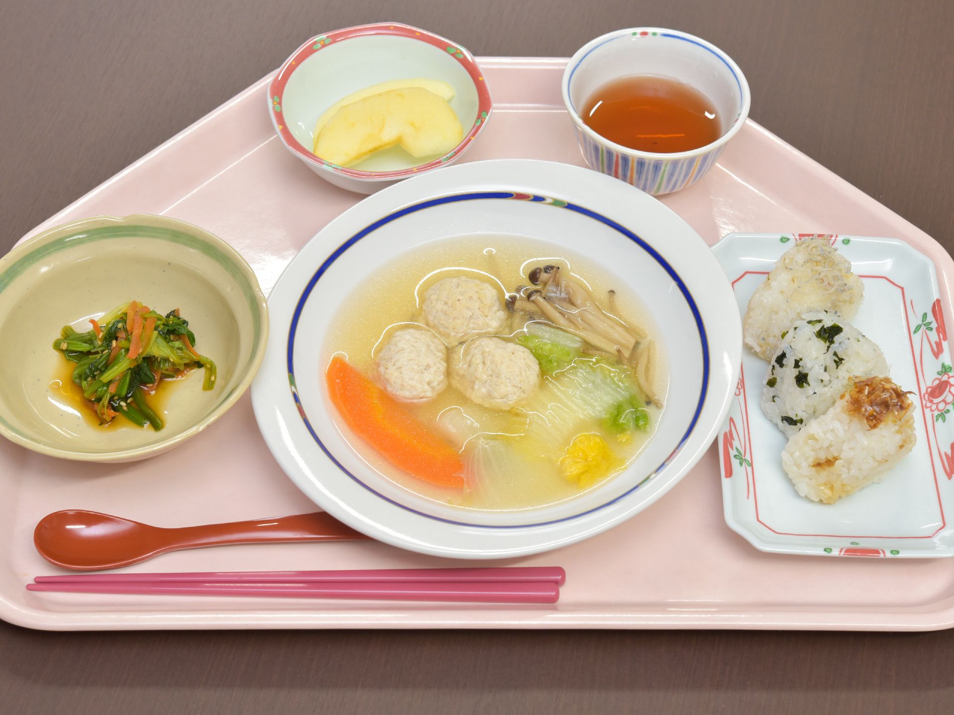 ピンクのトレーにはおにぎりが３つ、肉団子のスープ、野菜の和え物、リンゴ、お茶などが並んでいる。