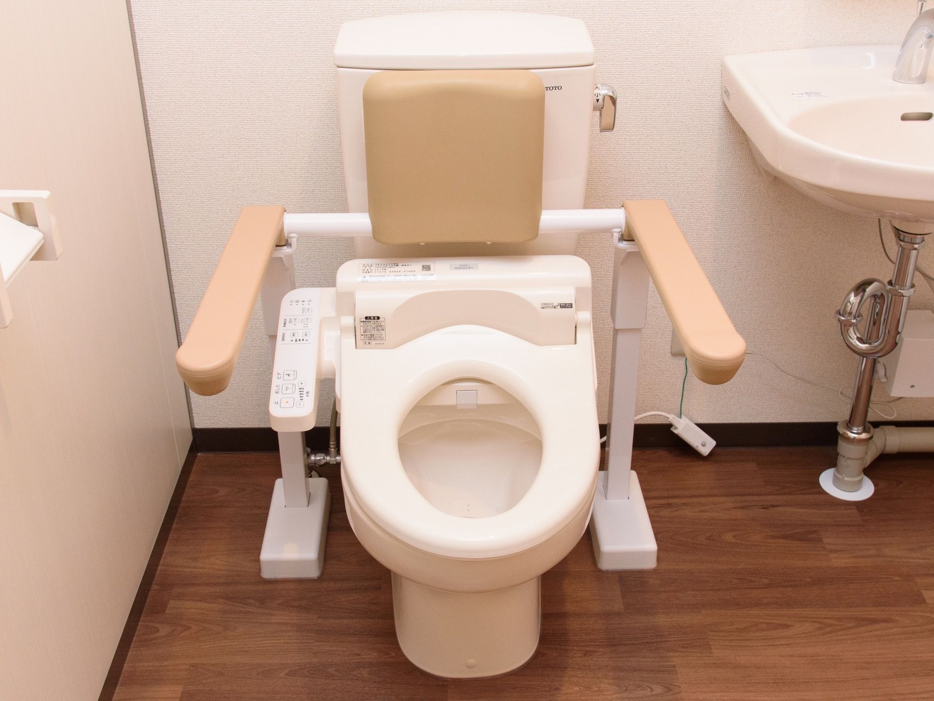 可動式の背もたれとひじ掛けが設置されたトイレである。清掃が行き届いたきれいなトイレになっており、隣に洗面台がある。