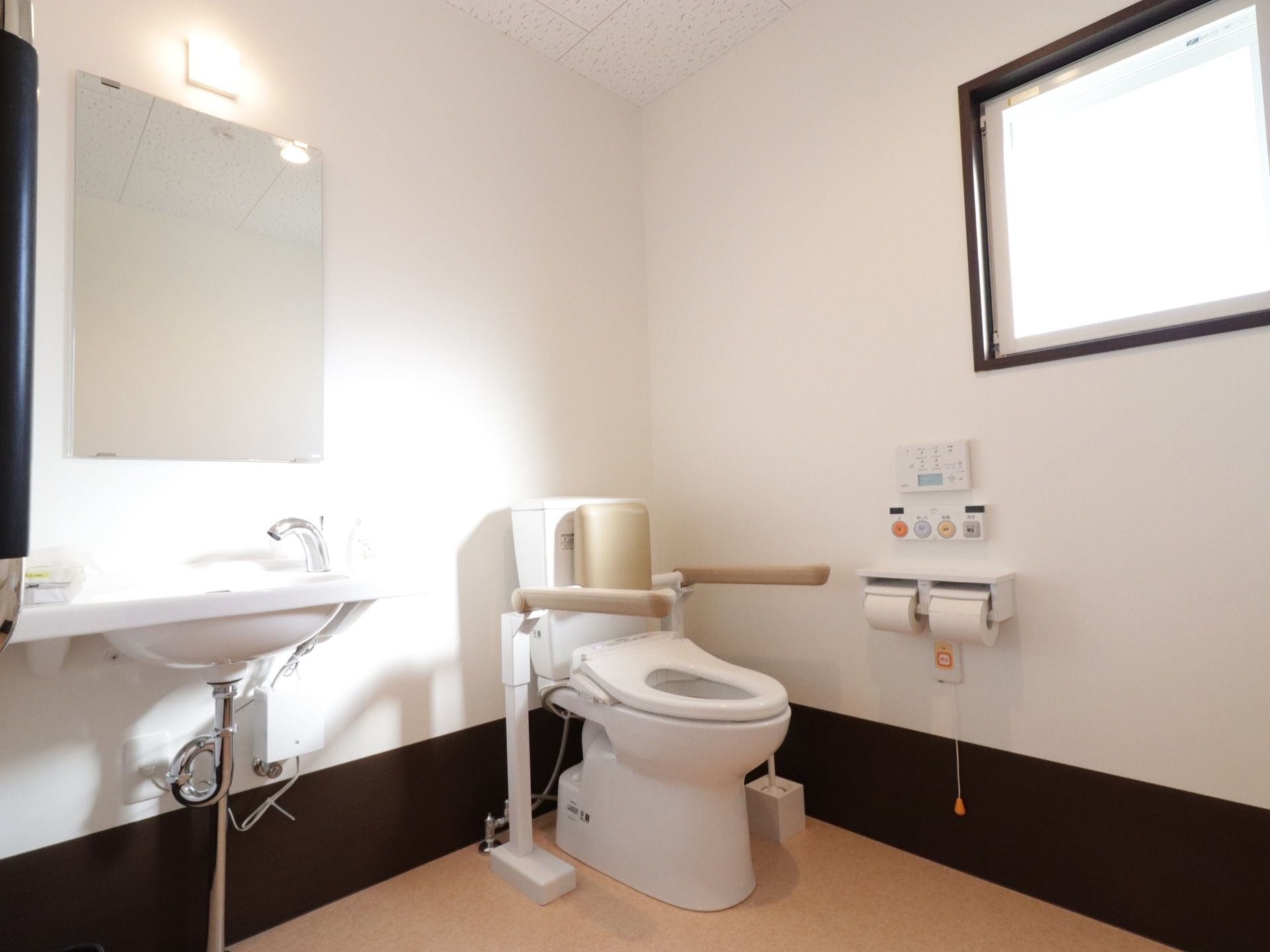 トイレには洗面台と洋式トイレが設置されている。トイレは背あてとひじ掛けが追加されている。壁には操作パネル、手すり、ナースコールが付いている。