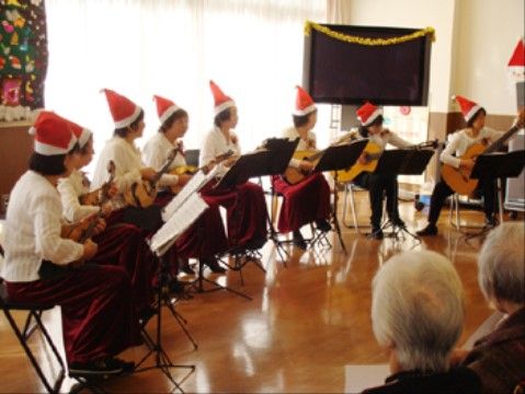 赤いクリスマスの帽子をかぶり、ギターを弾いている女性たちを眺めている入居者たち