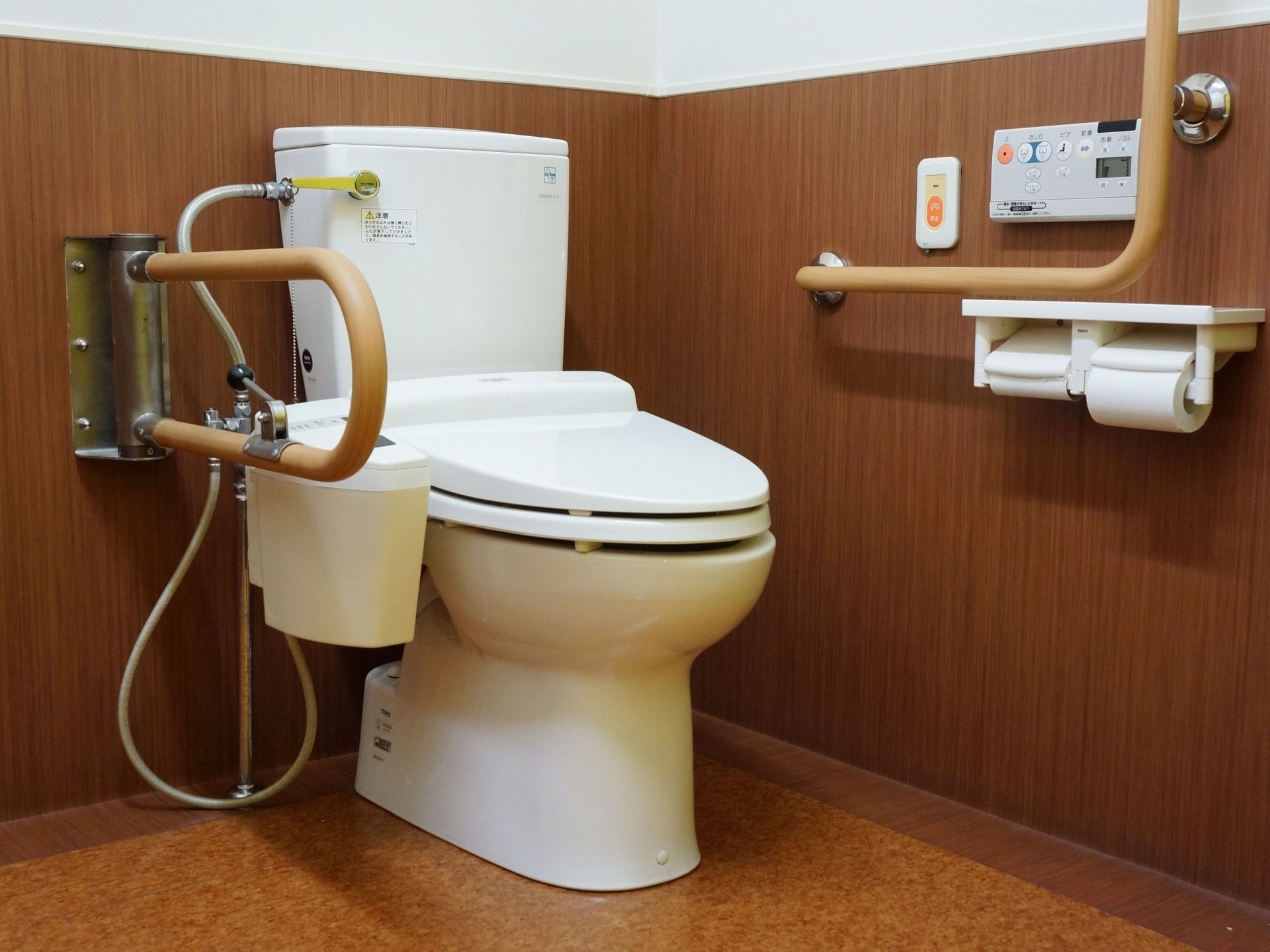 木目の壁がアクセントのトイレ。温水洗浄付きで、横に緊急通報ボタンが付いているので安心。手すりが左右に取り付けられ、立ち座りに優しい。