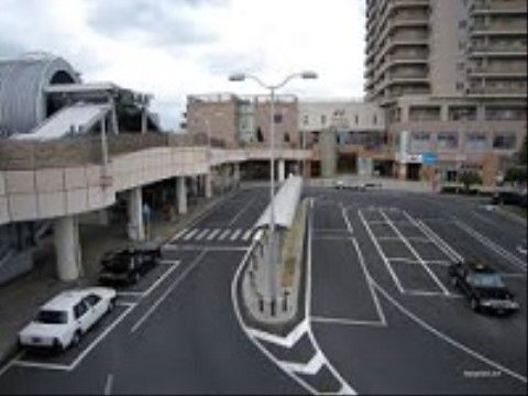 ロータリーのような広い道路で、駐車スペースに２台の車が停まっている。周辺にはさまざまな建物が立ち並んでいる。