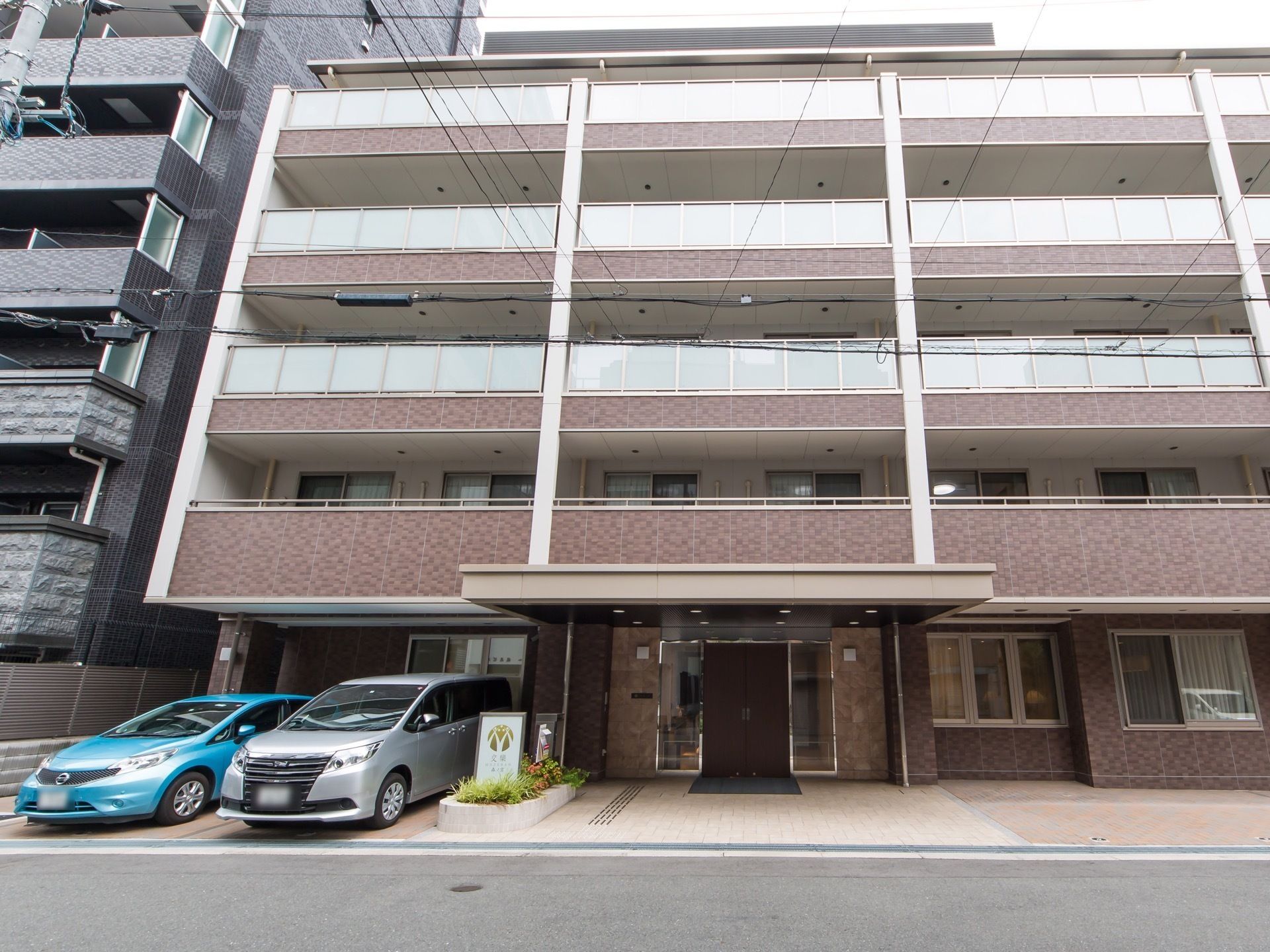 茶色を基調とした外壁の４階建ての施設である。玄関前には大きなひさしがあり、その左側に車が２台停まっている。