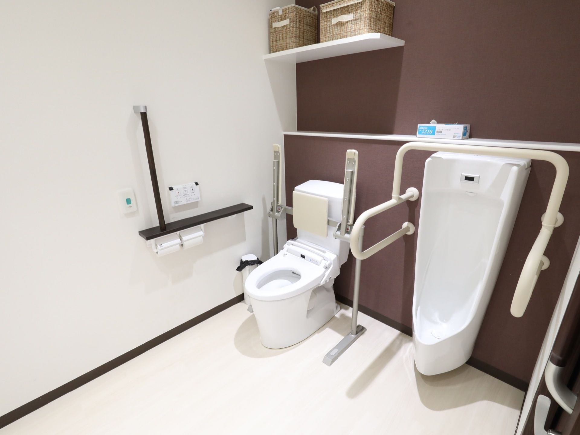 温水洗浄便座には折り畳み式の手すりと背もたれが付いている。隣には男性用便器が設置され、まわりには白い手すりがある。