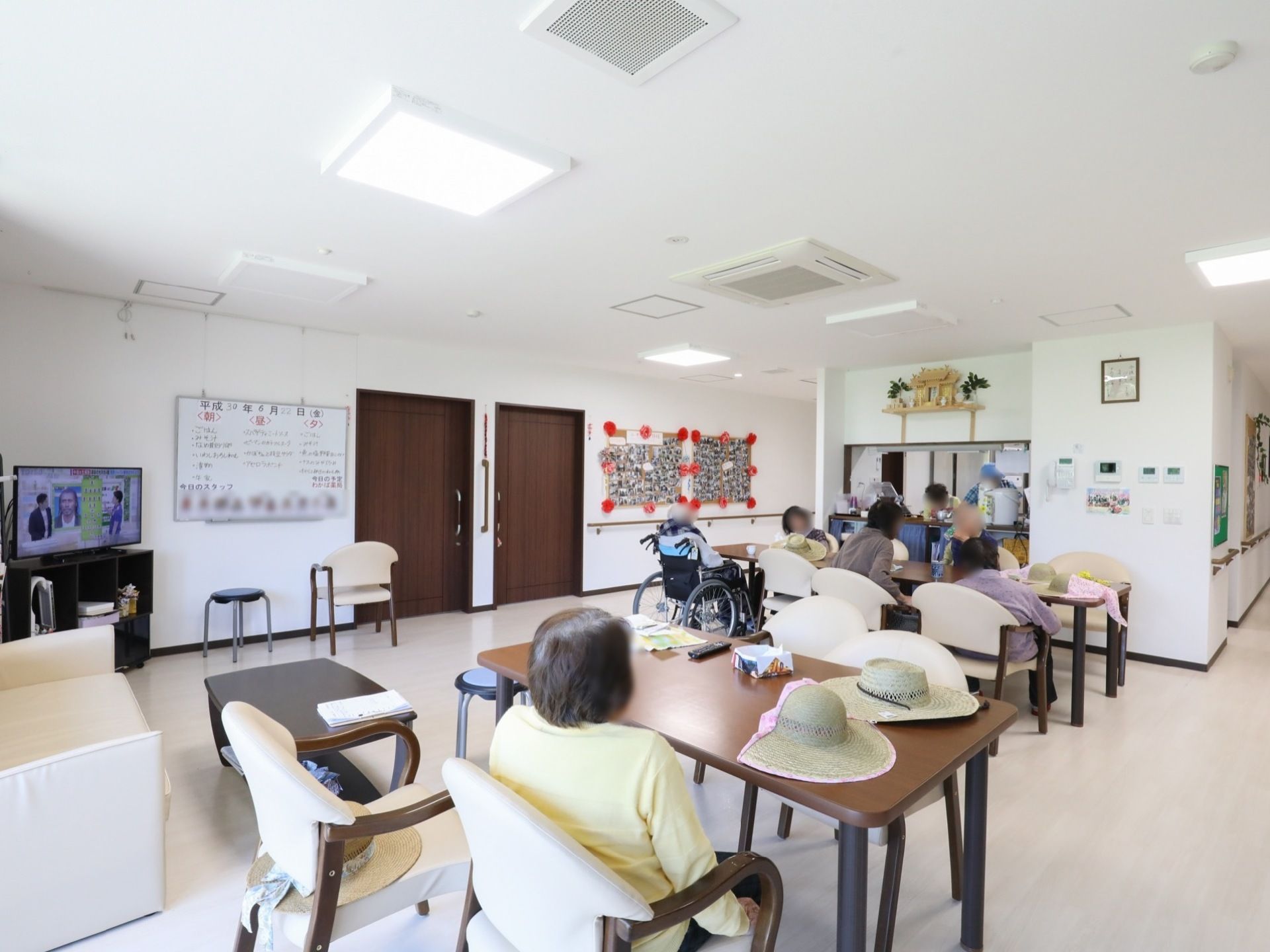 茶色のダイニングテーブルと白い椅子が設置され、入居者様が席についている。壁には大きなホワイトボードが掛けられ、献立が書かれている。