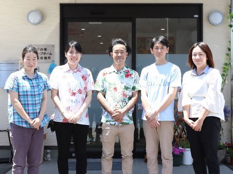 施設の玄関の前でほほえんでいる、アロハシャツなどを着た五人のスタッフ
