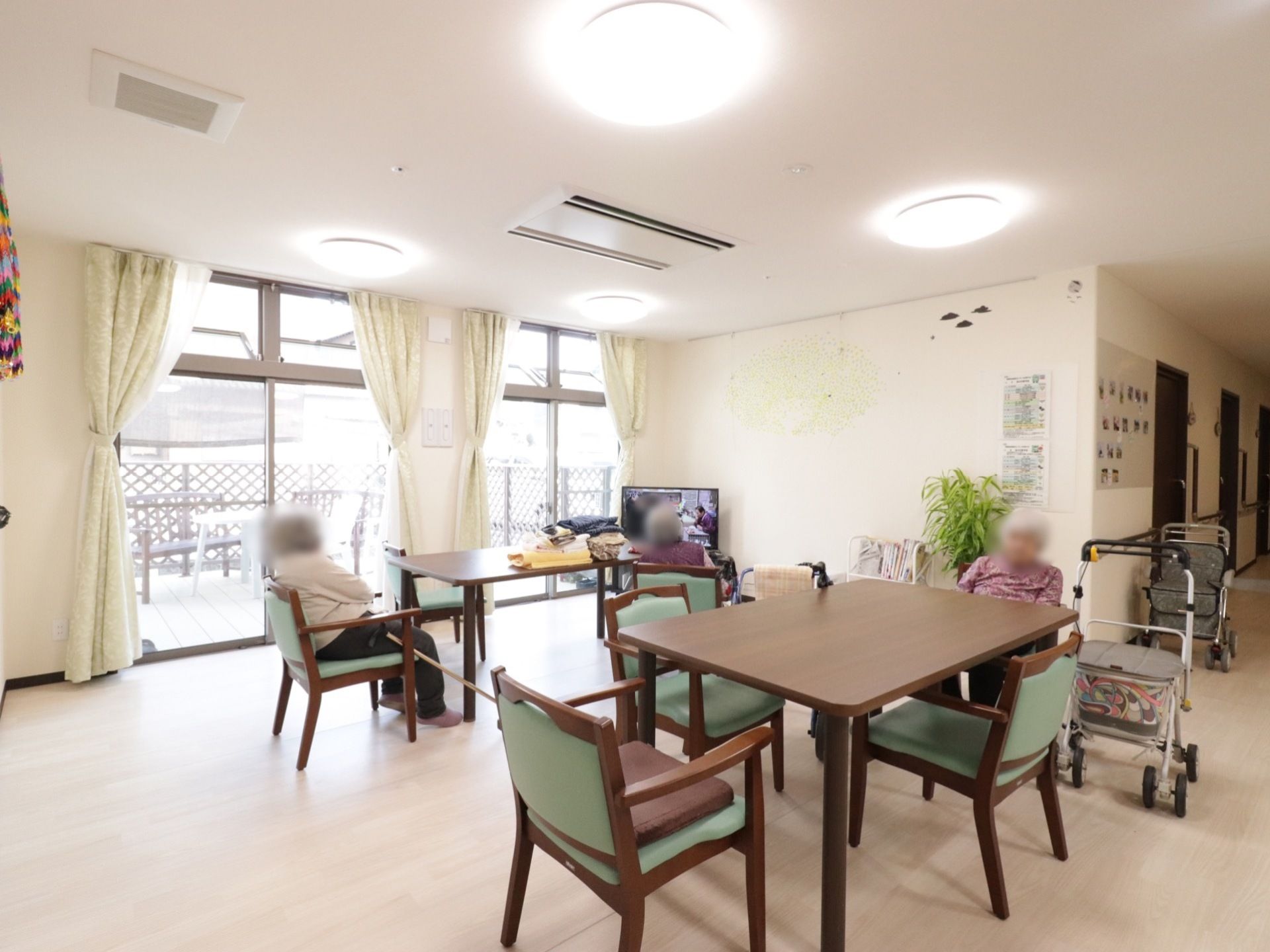 部屋の左奥に大きな窓があり、外にウッドデッキが見える。部屋の角にテレビが設置されていて、観葉植物も置かれている。
