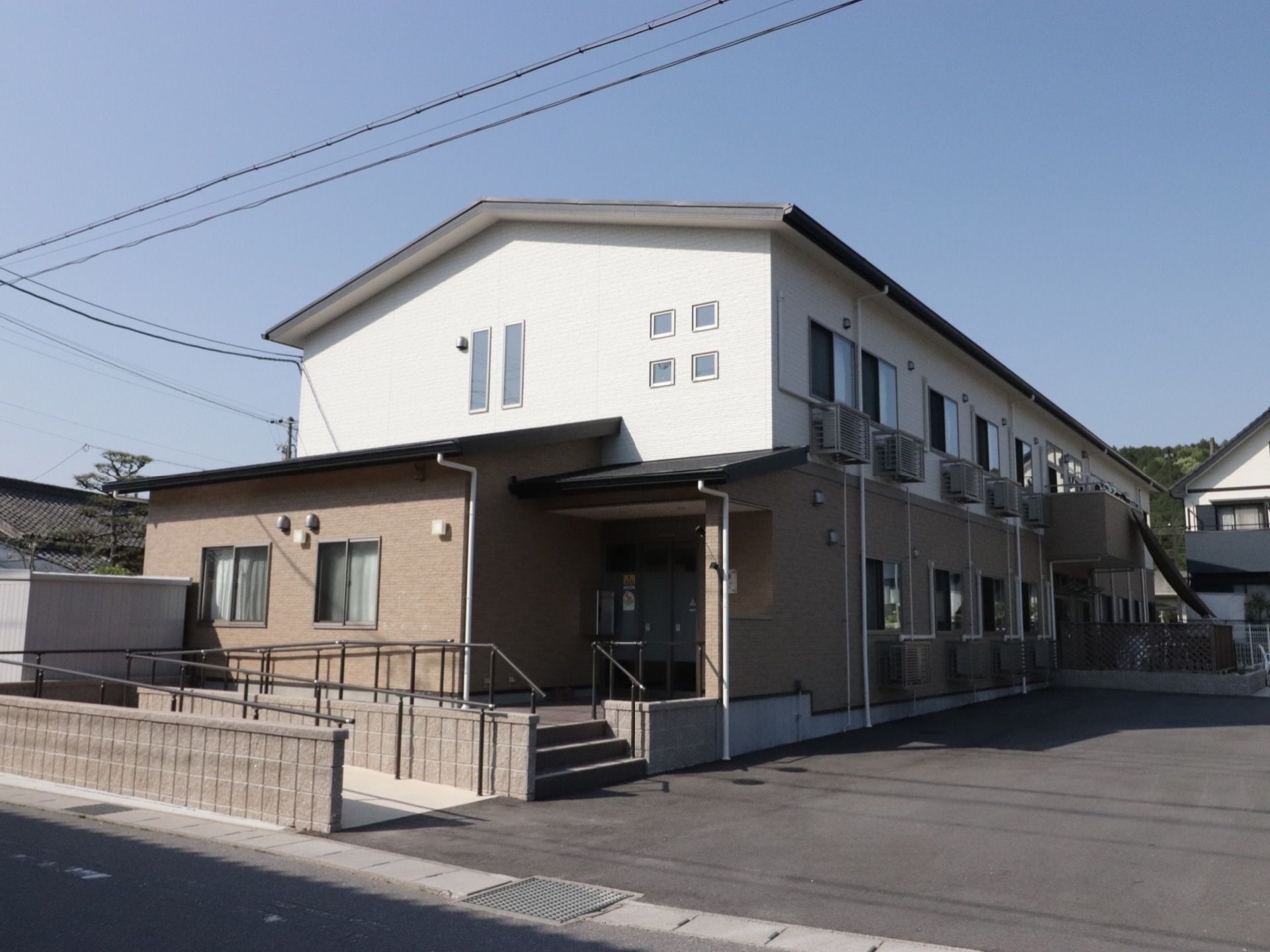 施設の建物は2階建てで、道路から縦に伸びている。手前にスロープがあり、玄関に続いている。建物の右手にコンクリートの広いスペースがある。