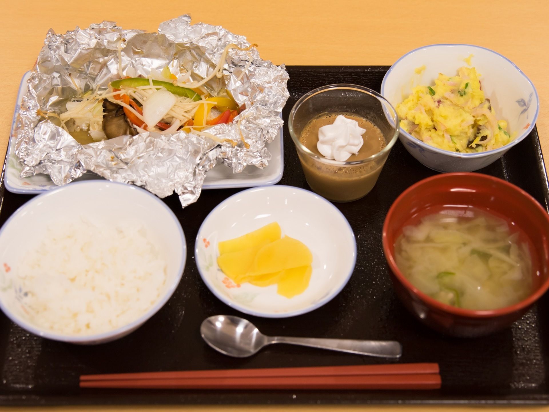 黒いお盆の上にご飯やお味噌汁、おかずなどが配膳されている。ホイル焼きにはいろいろな色の野菜が使用され、サラダも野菜が使用されている。