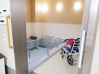 右側には浴室用の車椅子が置かれている。浴室内の壁の下半分は白色であり、上半分には木の板が張られている。