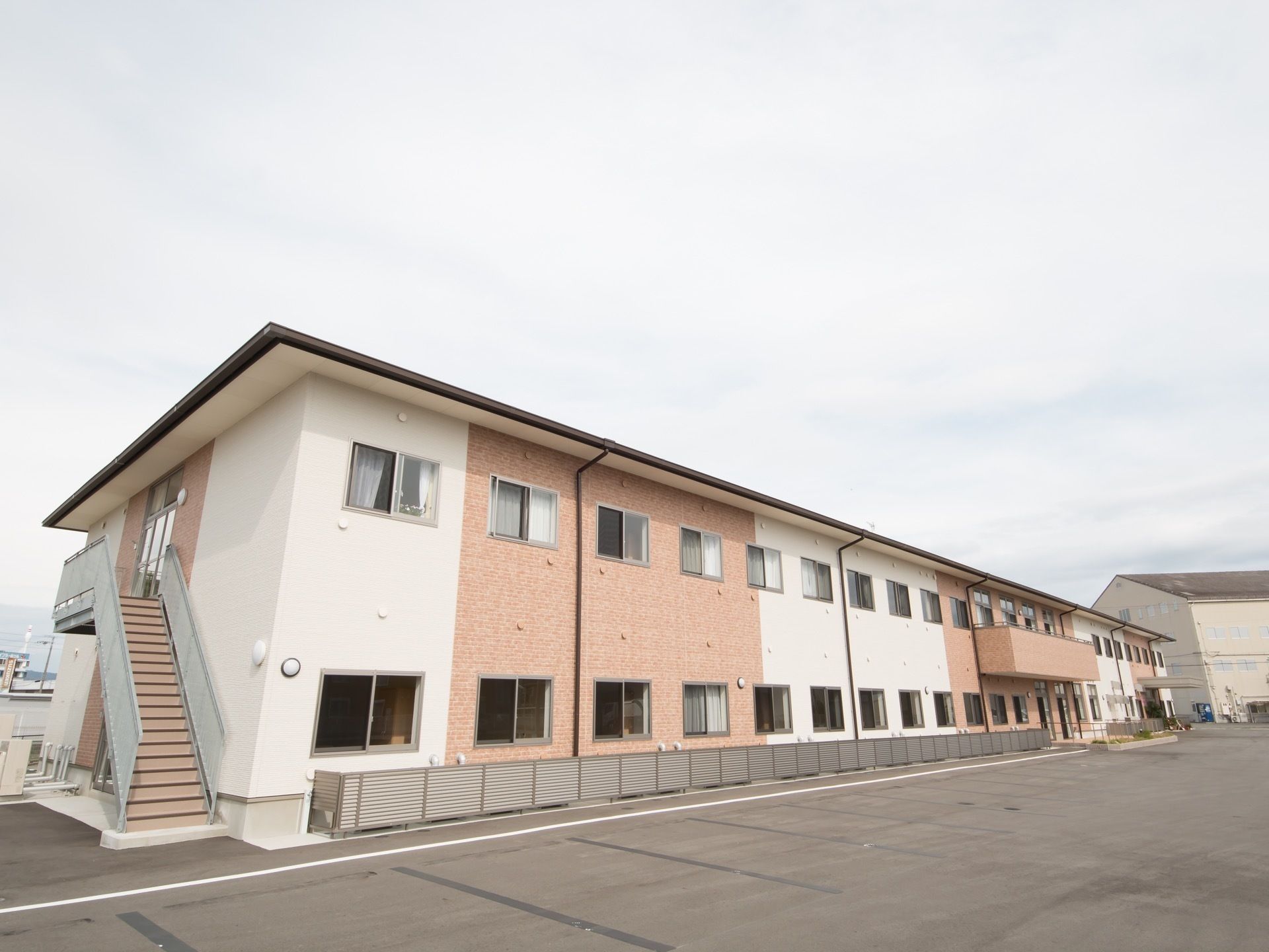 落ち着いた色合いに施された、2階建ての施設である。広々とした駐車スペースも用意されている。非常用の外階段も備えつけられている。