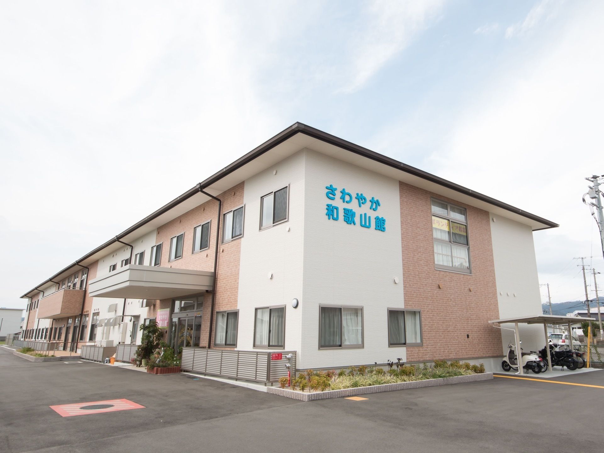 白とベージュ調の建物になっている。施設横には駐輪スペースが設けられており、バイクと自転車が停められている。