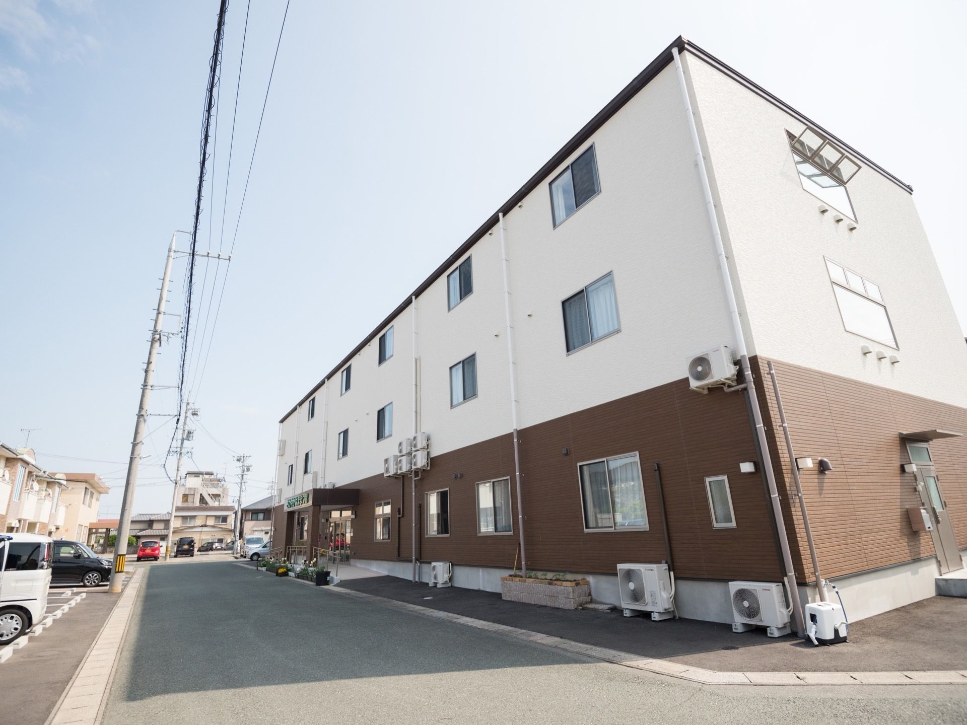 ブラウンを基調とした３階建ての落ち着いた色合いのグループホームで、周りにはエアコンの室外機が複数設置されている。