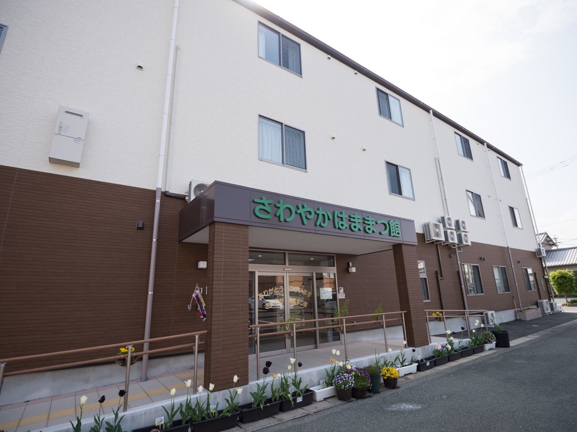 手すり付きのスロープが設置されている玄関で、手前には色とりどりの花が植えられたプランターや鉢植えが並べられている。