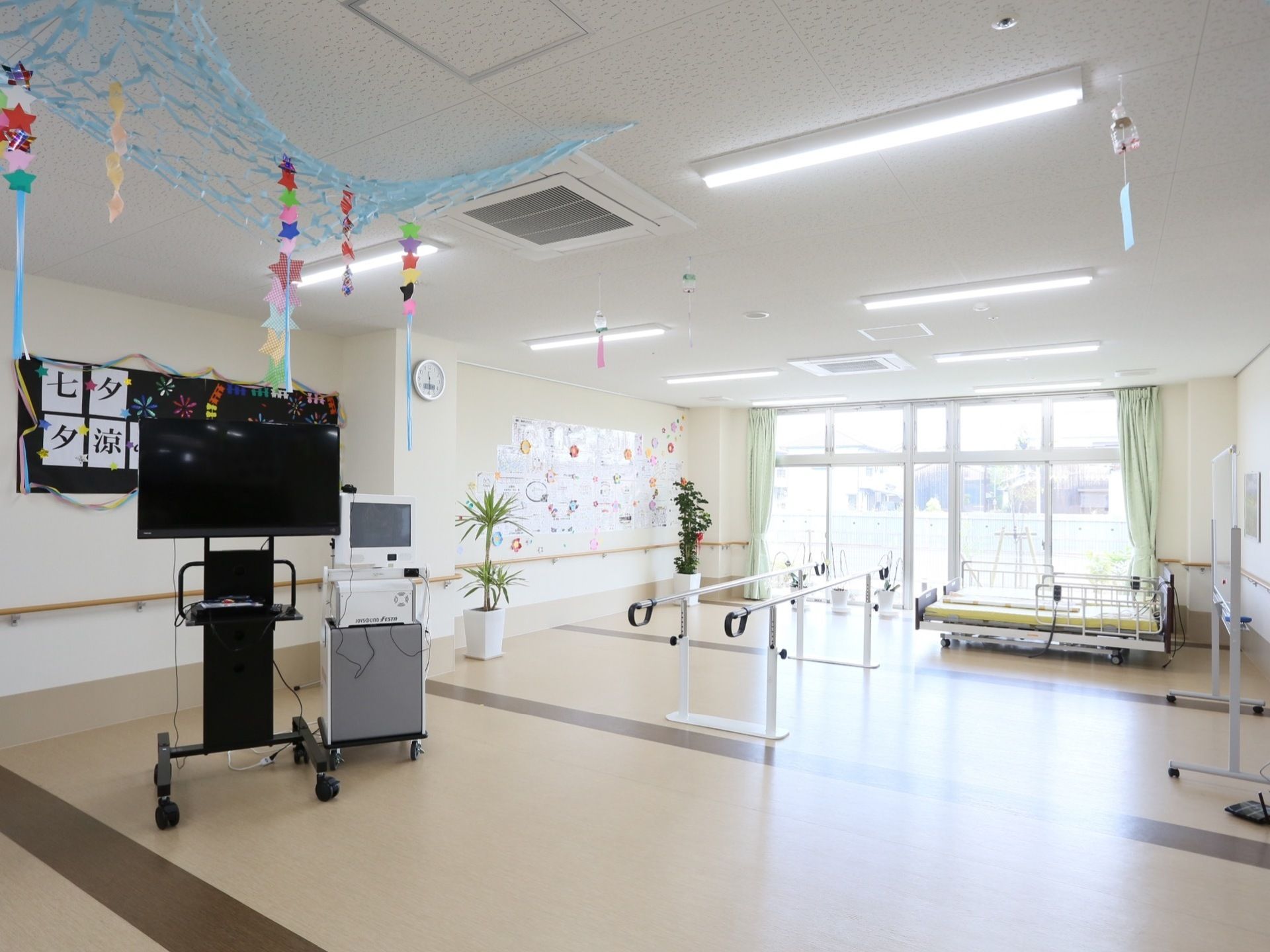 中央には平行棒が置いてあり、少し離れたところにキャスター付きの棚に乗った機械がある。窓の前には、電動ベッドがある。