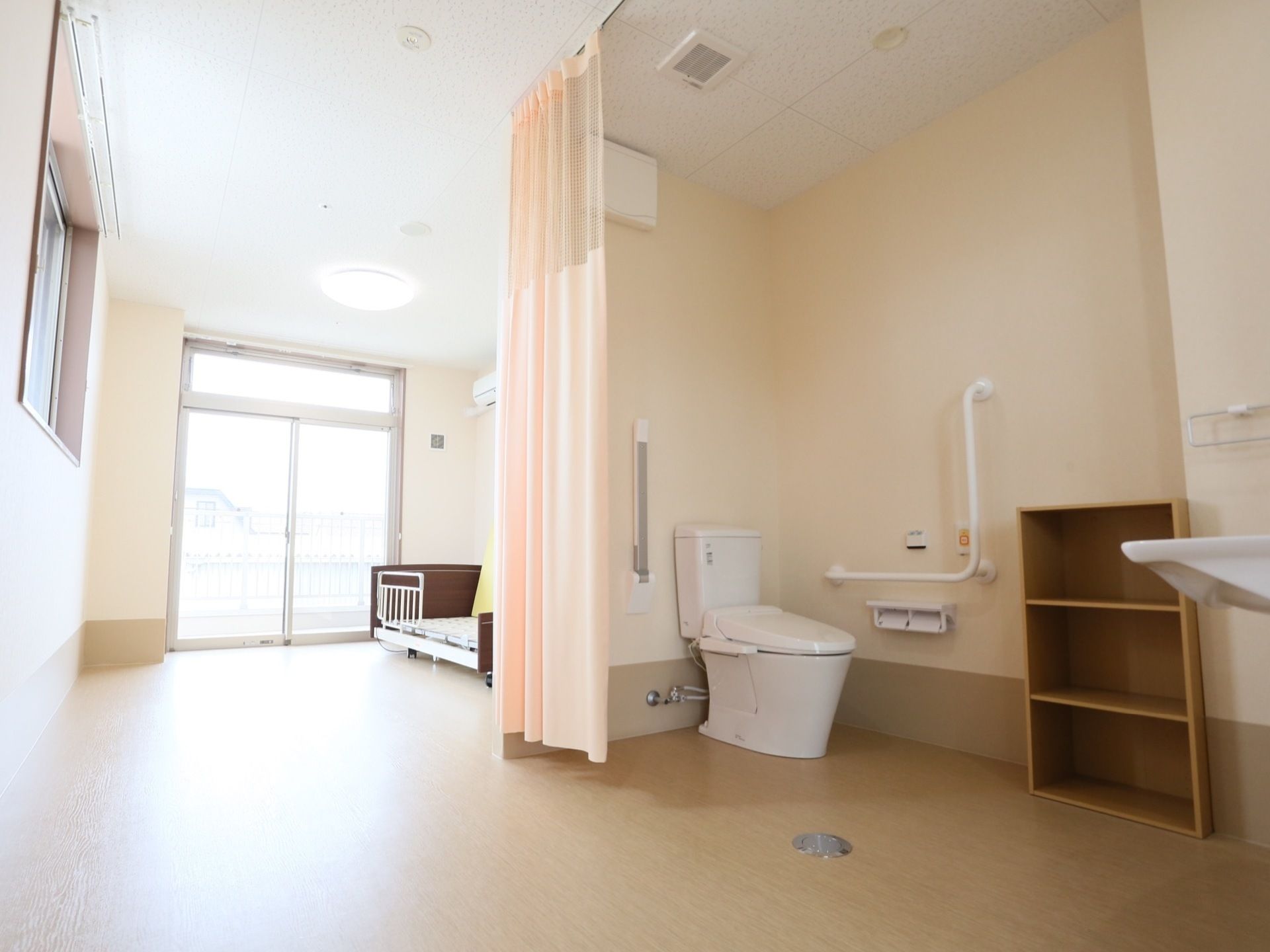 居室の一角に便座があり、カーテンで区切るようになっている。壁にはL字の手すりや緊急通報装置がついている。近くには木製の棚が置いてある。