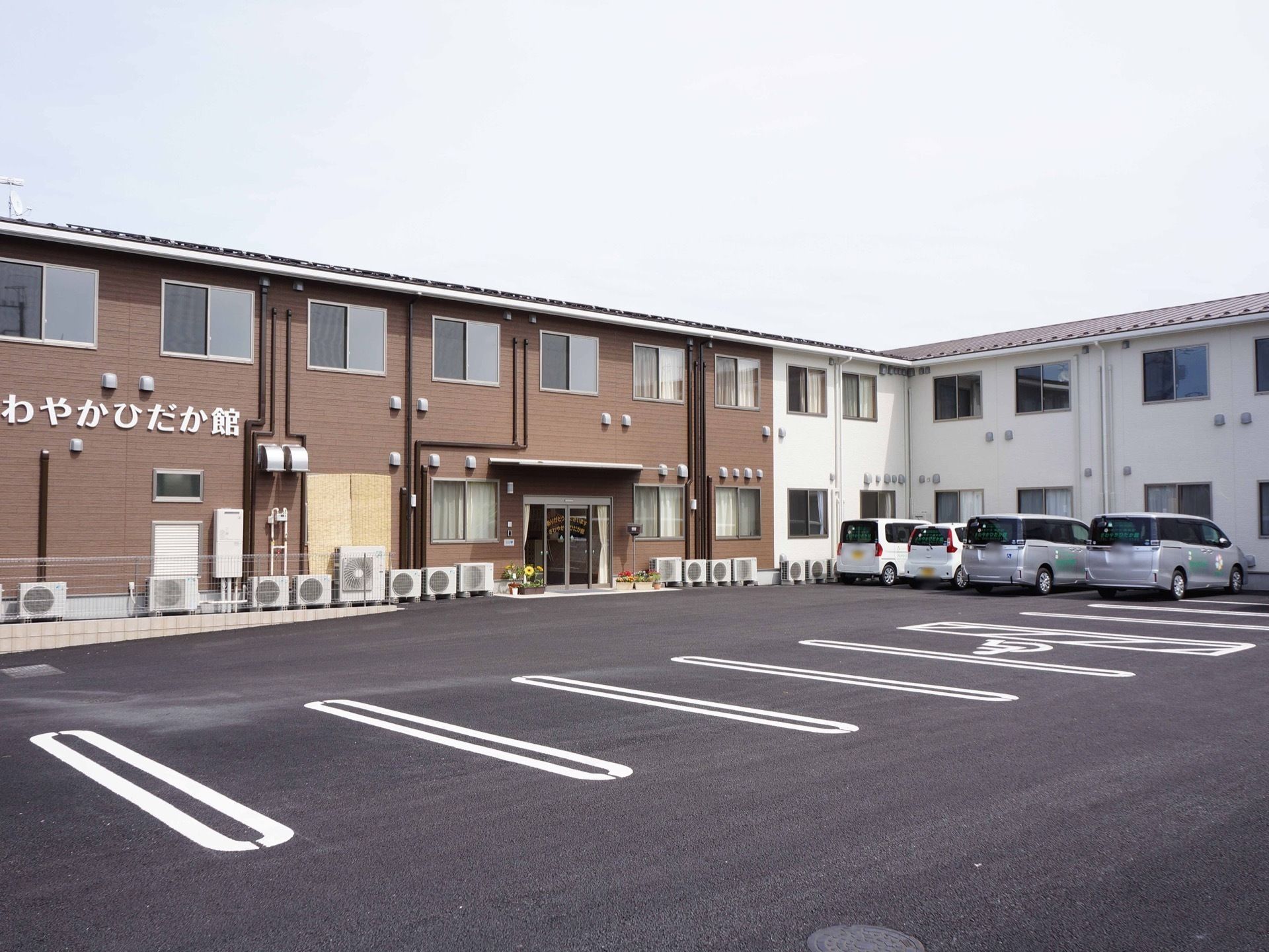 茶色と白の２色で施された、Ｌ字形の建物である。コンクリートの敷地に駐車場が完備され、幅にゆとりをもって駐車できる。