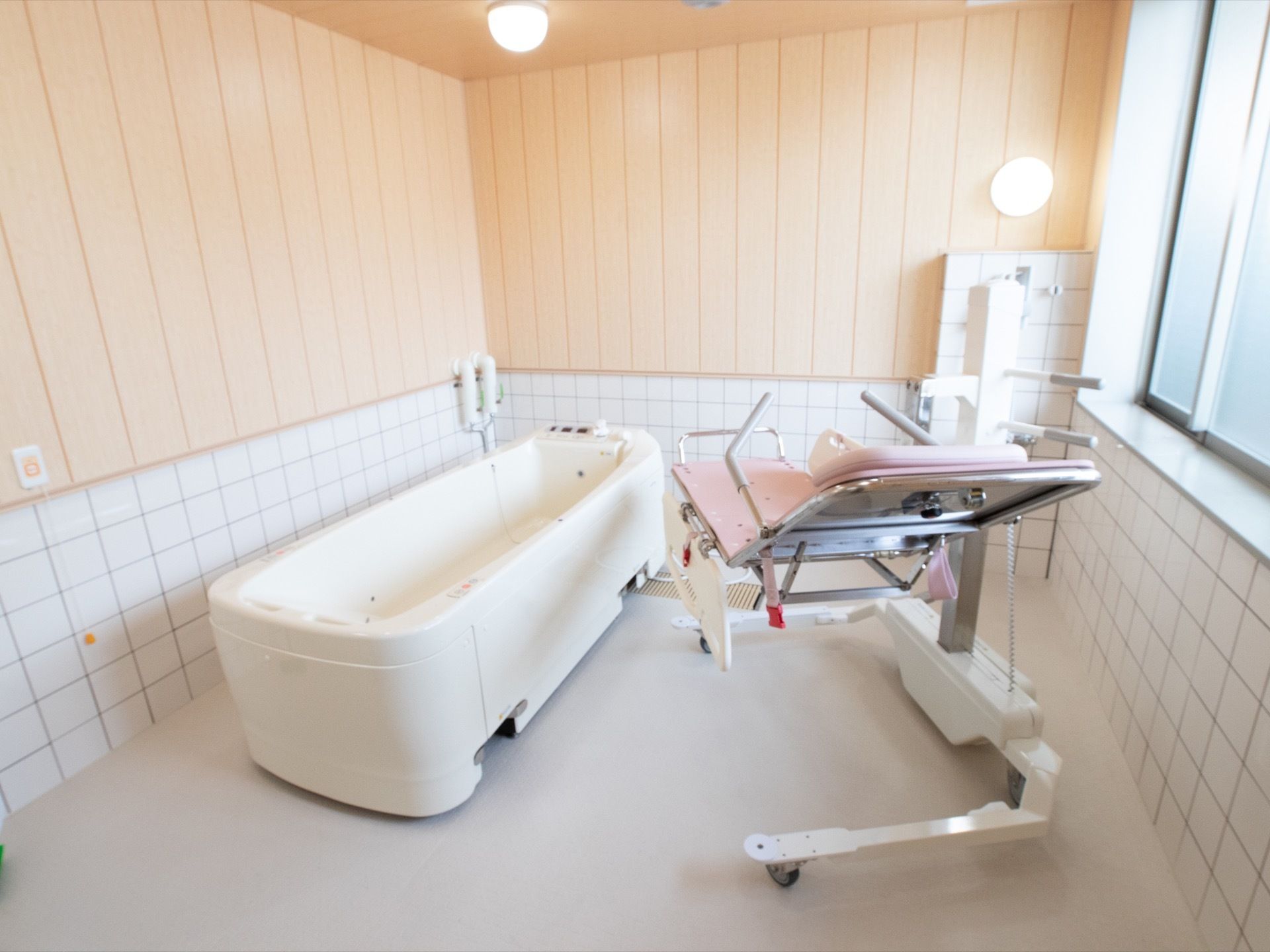 木の温もり感があふれる、広い浴室である。窓も設置されている。介護専用浴槽やピンク色のリフターが備え付けられている。