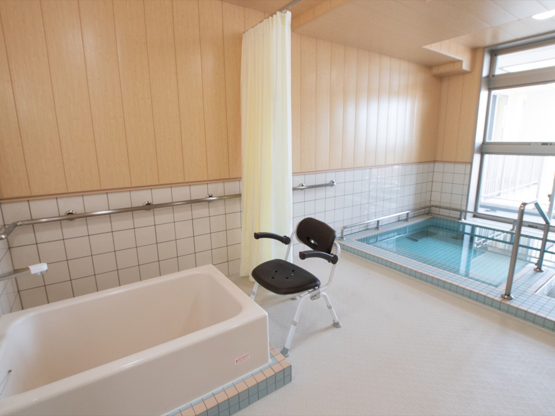 大きな窓の前に共用の浴槽がある。またカーテンで仕切り、一人で入浴できる浴槽とシャワーチェアも設置している。