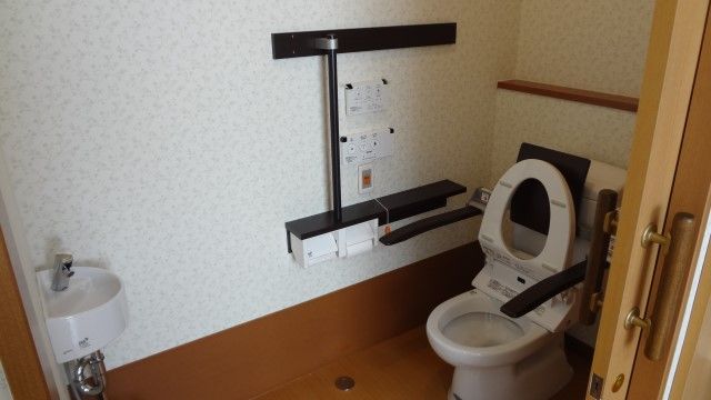 介護仕様のトイレで手すりや背もたれが取り付けられている。個室内に手洗い場も設けられている。壁にはナースコールボタンもある。