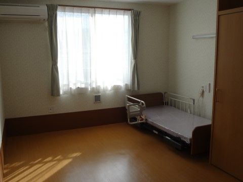 ゆったりとしたスペースのある居室には、ベッドが設置されている。他にエアコンや窓がある。床はフローリング材を使用。