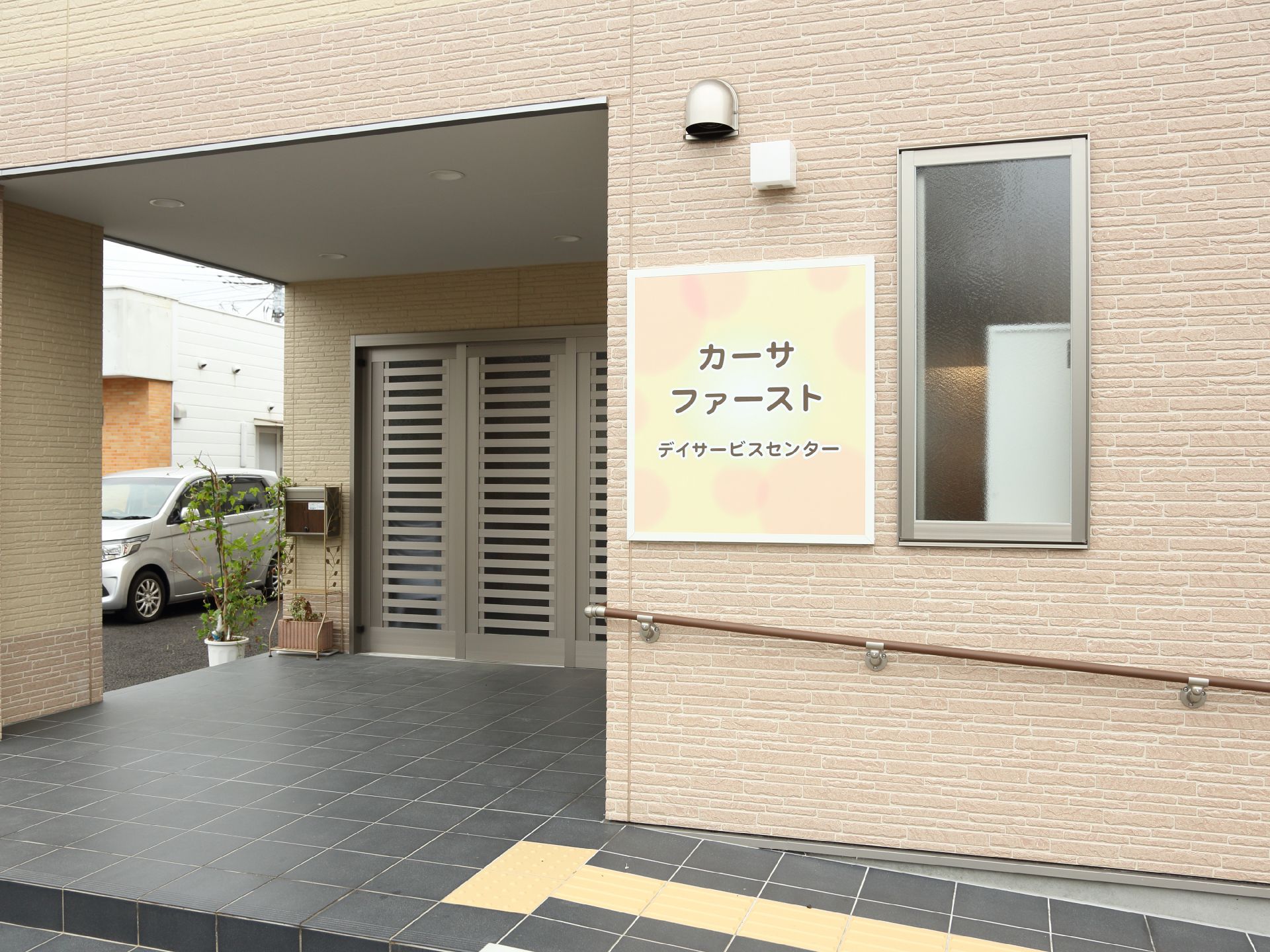 玄関前にはなだらかなスロープと手すりがついているので、車椅子のまま入室がスムーズにできる。玄関わきに車が一台停まっている。