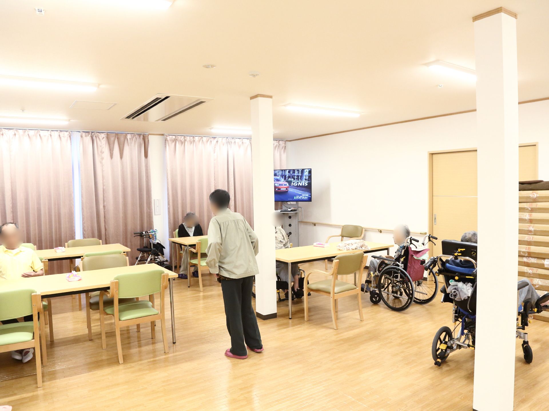 テーブルが四台あり、椅子が並べてある。椅子に入居者様が座っている。車椅子のまま席についている入居者様もいる。