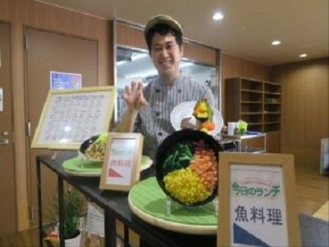 装飾台の上に今日のランチの見本が飾られており、その後ろで笑顔の男性が立っている様子