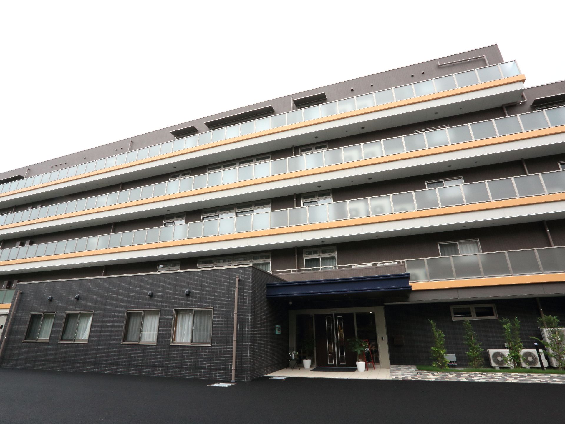 施設の外観は、壁がダークグレー色の1階中央分部に玄関があり、階上は半透明のベランダフェンス越しに各居室が並んでいるのが分かる。