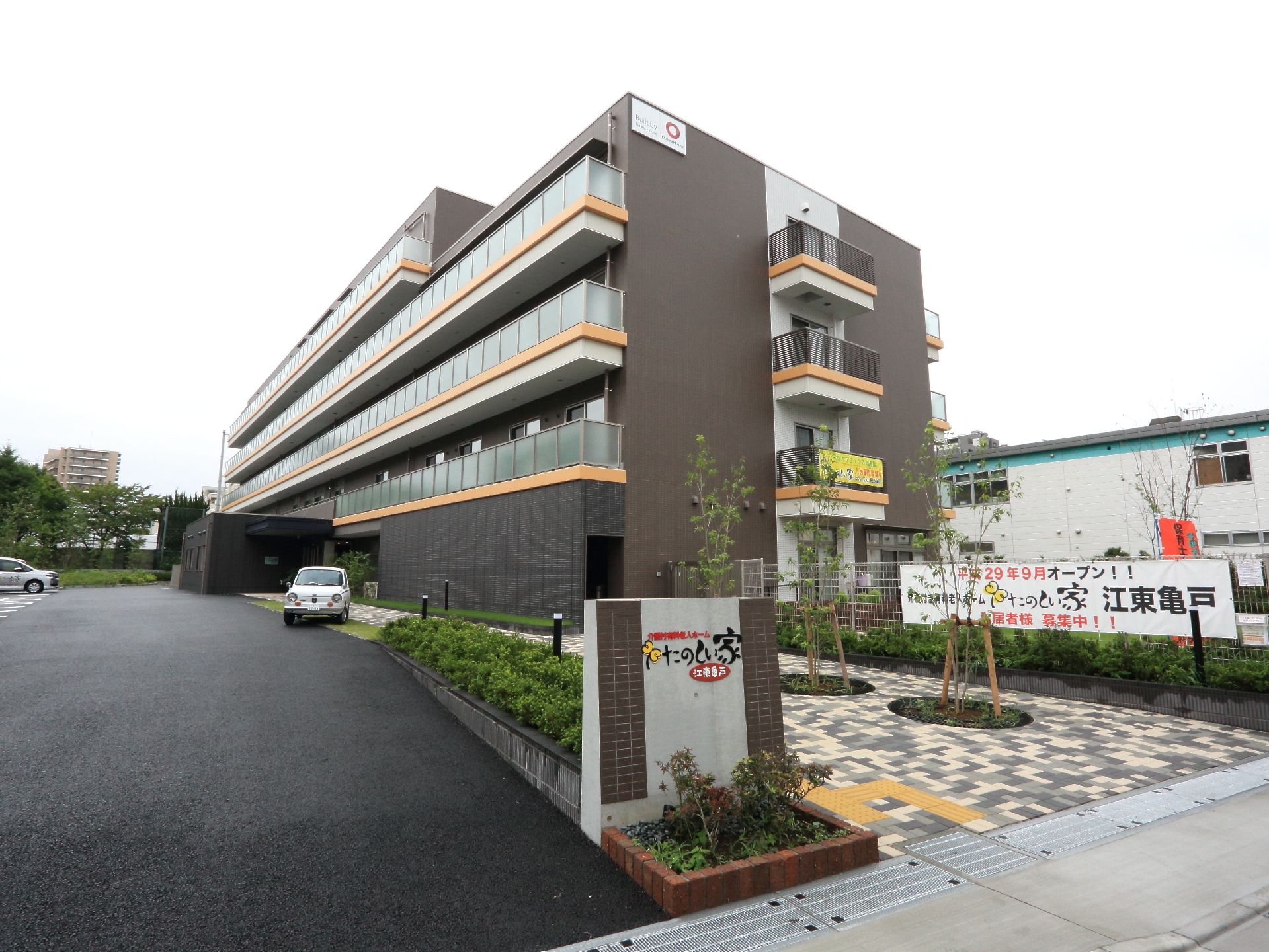 道路に面した門には施設名が書かれ所在地を示している。また門の横には、車道とは別に明るい色合いの歩道が玄関まで続いている。