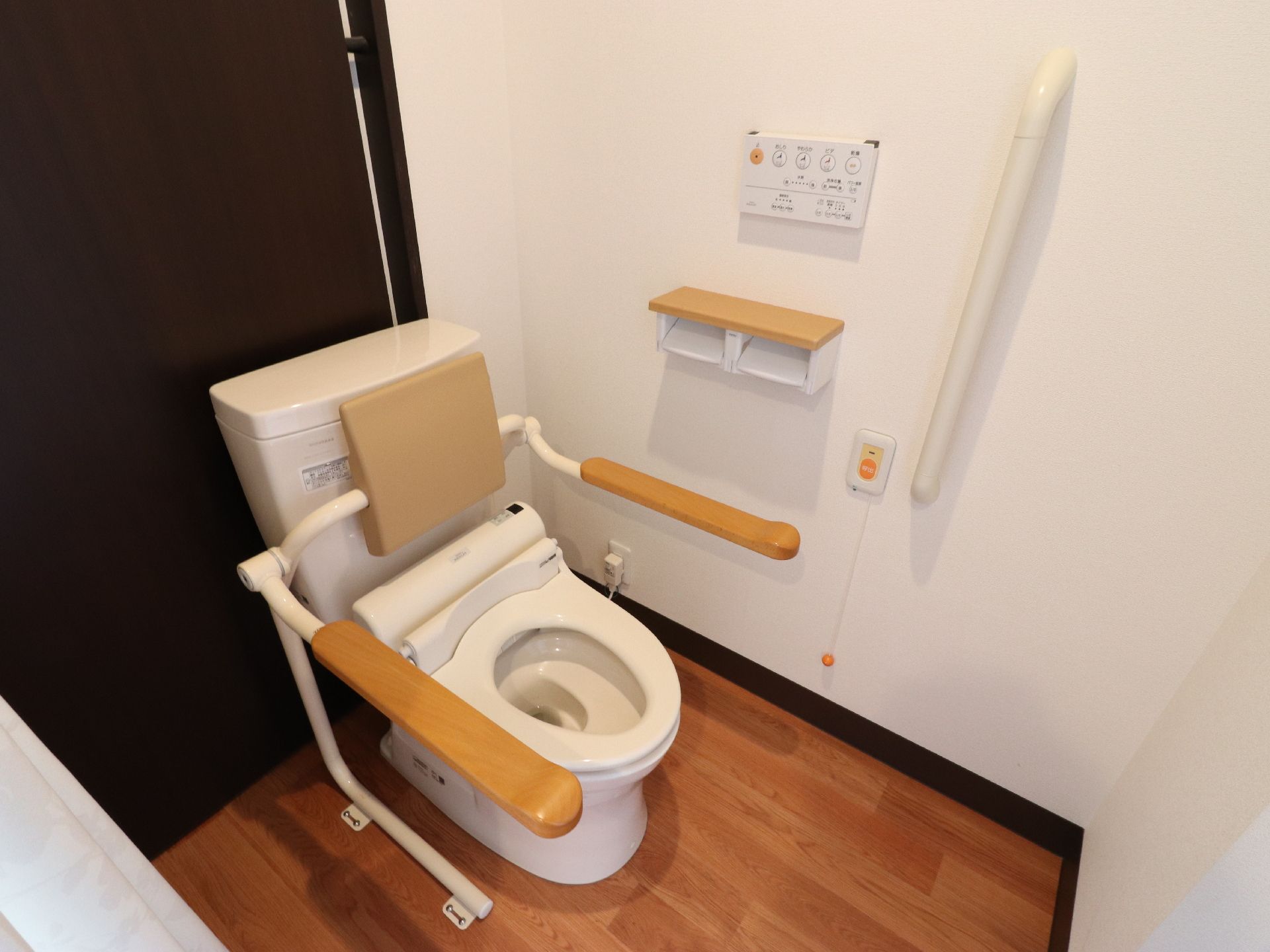 便器には背もたれと手すりが設置されている。背面は黒い壁になっており手すりのついた壁には呼び出しベルがついている。