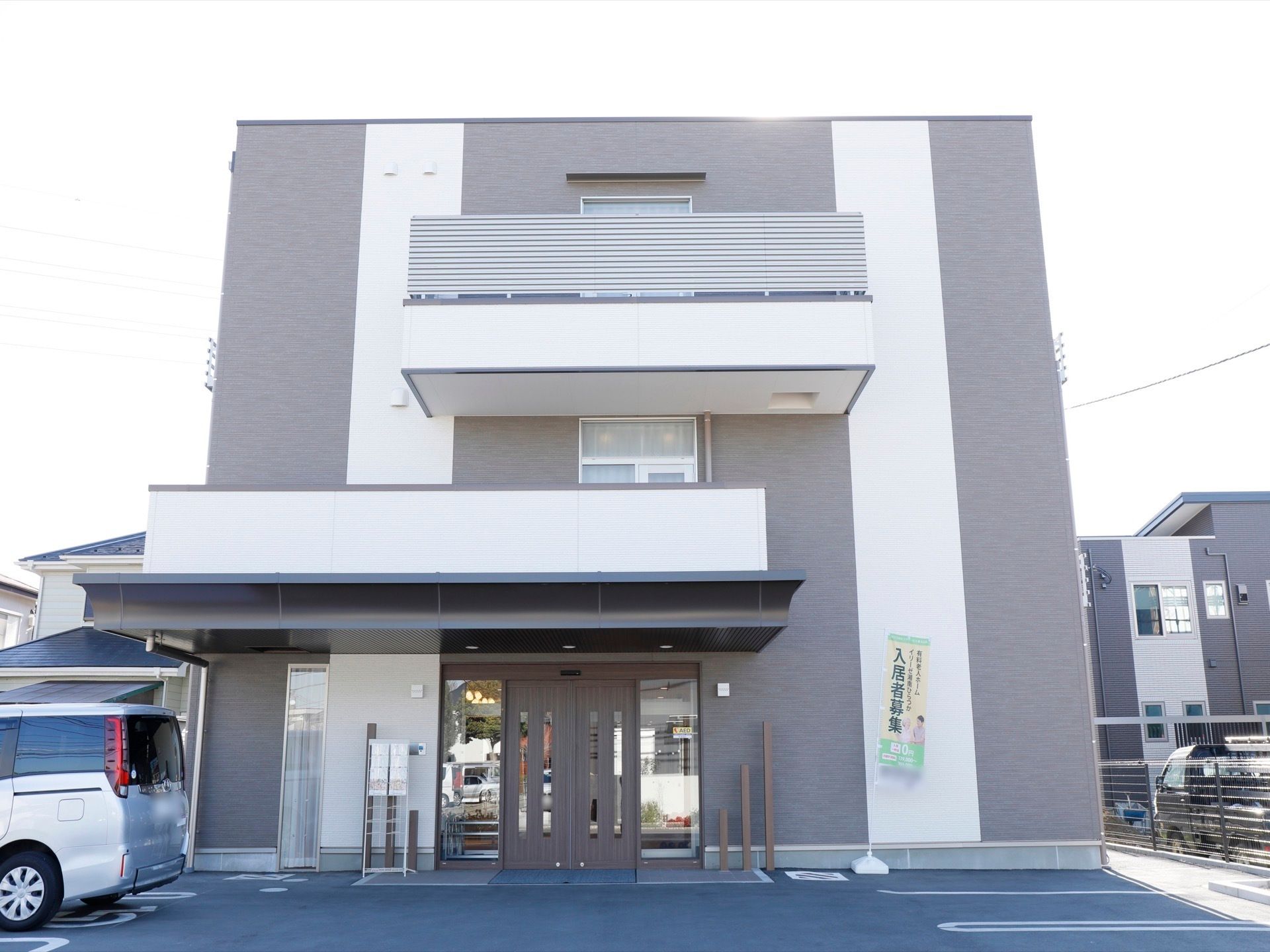 施設は３階建てで、灰色と白のシックなデザインである。駐車場からの段差は一つもなく、扉の右側には「入居者募集中」ののぼりが立っている。