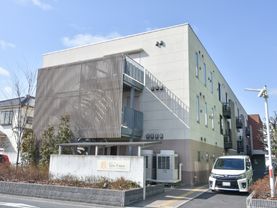 四角い白と茶に塗り分けられた施設になっている。外階段は装飾されており手前の花壇に施設名の入った看板がでている。