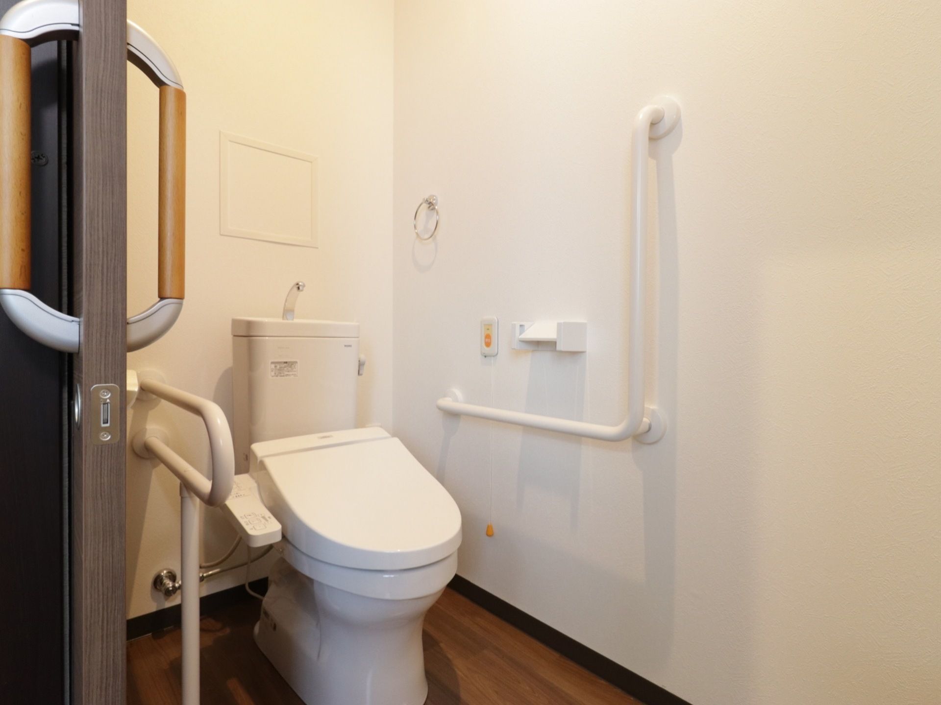 部屋の中にトイレがあり、壁にはL字型の手すりが付いている。そばには緊急呼び出しボタンや温水便座洗浄の操作パネルがある。